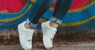 Person trägt weiße Sneaker und Jeans, steht vor bunter Wand.