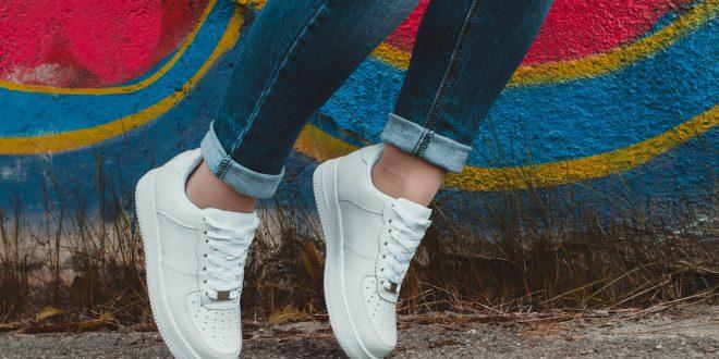 Person trägt weiße Sneaker und Jeans, steht vor bunter Wand.